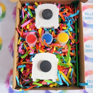 May include: A craft kit featuring two white ceramic bus shapes, three small paint pots in red, blue, and yellow, a green paintbrush, and colorful shredded paper in a brown cardboard box.