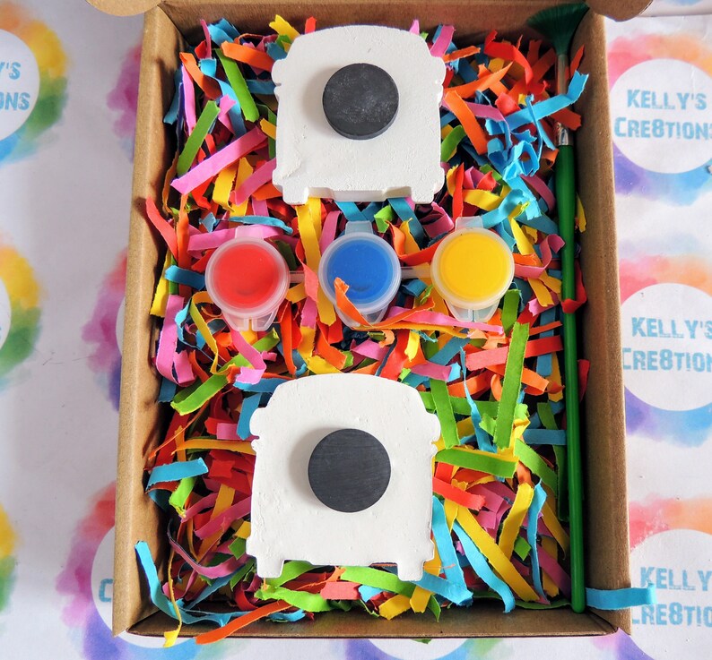 May include: A craft kit featuring two white ceramic bus shapes, three small paint pots in red, blue, and yellow, a green paintbrush, and colorful shredded paper in a brown cardboard box.
