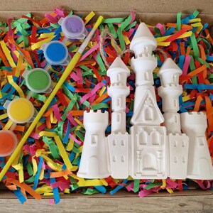 May include: A craft kit with a white castle figurine, paint pots in various colors, and a paintbrush. The kit is nestled in a box filled with colorful shredded paper.
