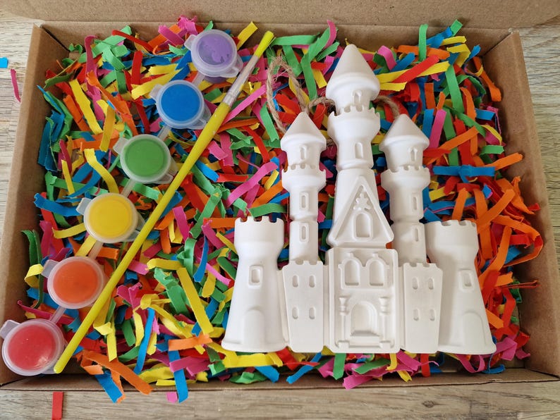 May include: A craft kit with a white castle figurine, paint pots in various colors, and a paintbrush. The kit is nestled in a box filled with colorful shredded paper.