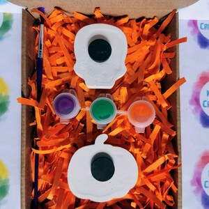 May include: A craft kit in a cardboard box, featuring a white skull and pumpkin, paint pots in purple, green, and orange, and a paintbrush. The box is filled with orange paper shreds. The text "KELLY'S CREATIONS" is visible.