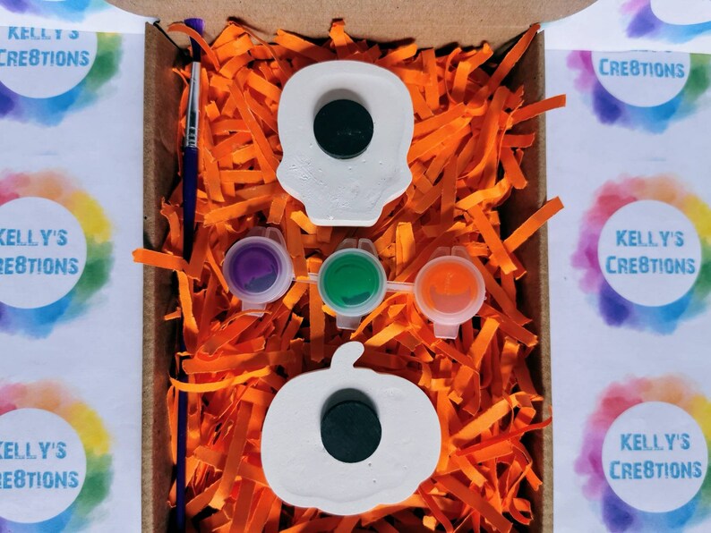 May include: A craft kit in a cardboard box, featuring a white skull and pumpkin, paint pots in purple, green, and orange, and a paintbrush. The box is filled with orange paper shreds. The text "KELLY'S CREATIONS" is visible.