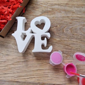 May include: White wooden LOVE sign with a heart cutout in the O. The sign is on a wooden surface with pink paint pots and a paintbrush.