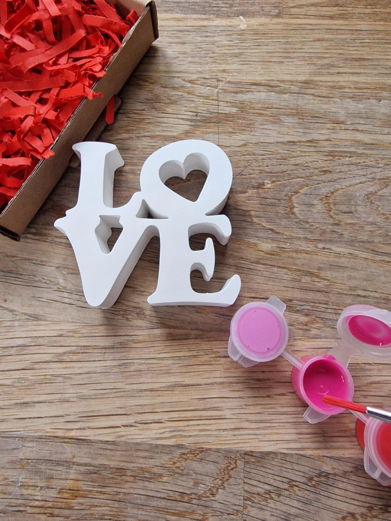 May include: White wooden LOVE sign with a heart cutout in the O. The sign is on a wooden surface with pink paint pots and a paintbrush.