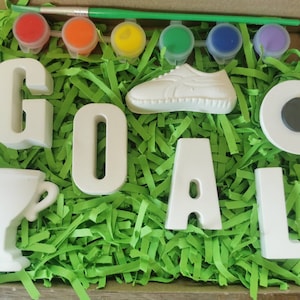 May include: A craft kit with white ceramic letters spelling "GOAL", a white ceramic trophy, a white ceramic shoe, and a white ceramic circle with a black circle in the center. The kit also includes six small paint pots with red, orange, yellow, green, blue, and purple paint.
