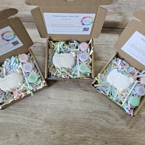 May include: Three open brown cardboard boxes containing a paint your own kit. Each kit includes a white ceramic cat figure, a small paintbrush, and three small round containers of pastel paint nestled in colourful shredded paper filler.