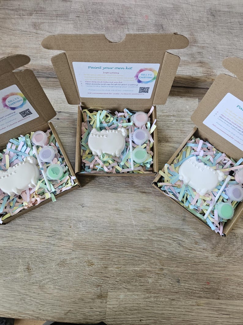 May include: Three open brown cardboard boxes containing a paint your own kit. Each kit includes a white ceramic cat figure, a small paintbrush, and three small round containers of pastel paint nestled in colourful shredded paper filler.