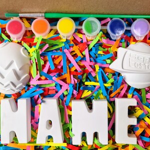 May include: A craft kit with white ceramic eggs, letters spelling "NAME", and paint pots in red, orange, yellow, green, blue, and purple. The eggs are decorated with a zig-zag pattern and the words "Happy Easter" are on one of the eggs. The kit is surrounded by colorful shredded paper.