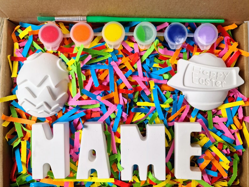 May include: A craft kit with white ceramic eggs, letters spelling "NAME", and paint pots in red, orange, yellow, green, blue, and purple. The eggs are decorated with a zig-zag pattern and the words "Happy Easter" are on one of the eggs. The kit is surrounded by colorful shredded paper.