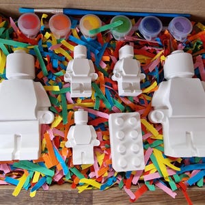 May include: A set of five white ceramic figurines in the shape of Lego people, a Lego brick, and five small pots of paint. The figurines and brick are in a box filled with colorful shredded paper.