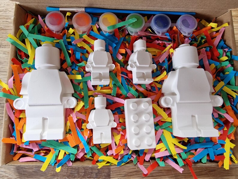 May include: A set of five white ceramic figurines in the shape of Lego people, a Lego brick, and five small pots of paint. The figurines and brick are in a box filled with colorful shredded paper.