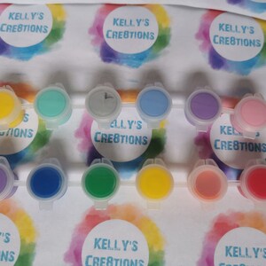 May include: A set of 10 small paint pots with lids. The pots are arranged in two rows of five. The paint colors are yellow, green, blue, purple, pink, gray, and red. The lids are white with the text "Kelly's Creations" printed on them.