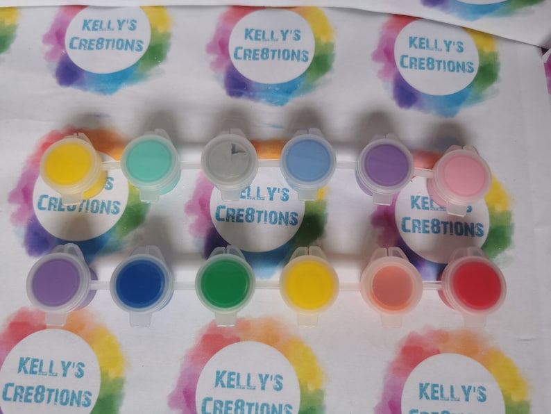 May include: A set of 10 small paint pots with lids. The pots are arranged in two rows of five. The paint colors are yellow, green, blue, purple, pink, gray, and red. The lids are white with the text "Kelly's Creations" printed on them.