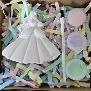 Op de afbeelding: Een wit keramieken prinsessenfiguur in een baljurk, een witte kwast en drie kleine ronde plastic bakjes gevuld met pastelroze, paarse en groene verf, allemaal in een doos gevuld met versnipperde stroken pastelkleurig papier.