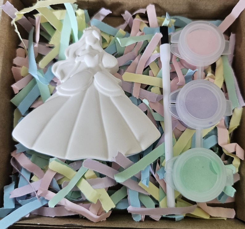 Op de afbeelding: Een wit keramieken prinsessenfiguur in een baljurk, een witte kwast en drie kleine ronde plastic bakjes gevuld met pastelroze, paarse en groene verf, allemaal in een doos gevuld met versnipperde stroken pastelkleurig papier.