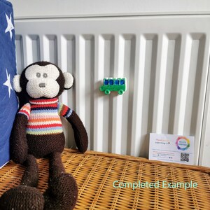May include: A brown knitted monkey toy sitting on a wicker basket. The monkey is wearing a striped sweater. A green toy bus is attached to a white radiator behind the monkey.