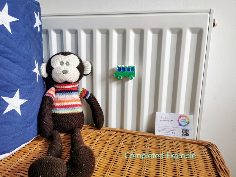 May include: A brown knitted monkey toy sitting on a wicker basket. The monkey is wearing a striped sweater. A green toy bus is attached to a white radiator behind the monkey.