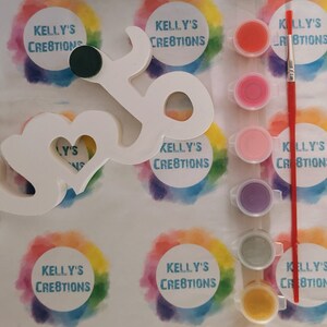 May include: A white wooden sign in the shape of the word "love" with a heart cutout in the middle. The sign is surrounded by paint pots and a paintbrush. The paint pots are in various colours, including red, pink, purple, green, and yellow. The sign is on a white background with rainbow circles that say "Kelly's Creations".
