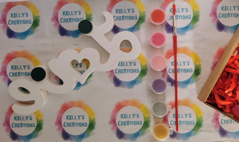 May include: A white wooden sign in the shape of the word "love" with a heart cutout in the middle. The sign is surrounded by paint pots and a paintbrush. The paint pots are in various colours, including red, pink, purple, green, and yellow. The sign is on a white background with rainbow circles that say "Kelly's Creations".