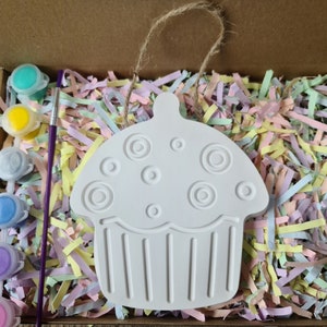May include: A white ceramic cupcake ornament with a swirl design and a hanging loop. The ornament is surrounded by colourful paper shreds and paint pots.