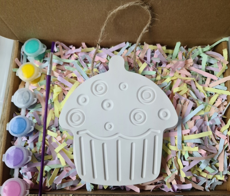 May include: A white ceramic cupcake ornament with a swirl design and a hanging loop. The ornament is surrounded by colourful paper shreds and paint pots.