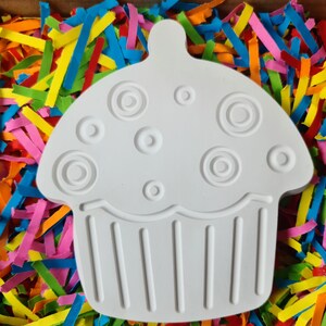 May include: White ceramic cupcake shaped ornament with a swirl design on top and a ribbed bottom. The ornament is surrounded by colourful paper shreds.