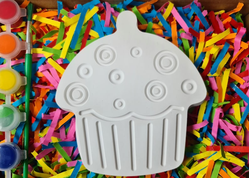 May include: White ceramic cupcake shaped ornament with a swirl design on top and a ribbed bottom. The ornament is surrounded by colourful paper shreds.