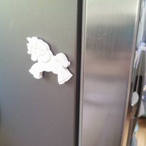 May include: A white, three-dimensional horse-shaped magnet is affixed to a dark gray refrigerator door. The magnet is a stylized depiction of a horse, with a flowing mane and tail. The refrigerator door is next to a stainless steel door.