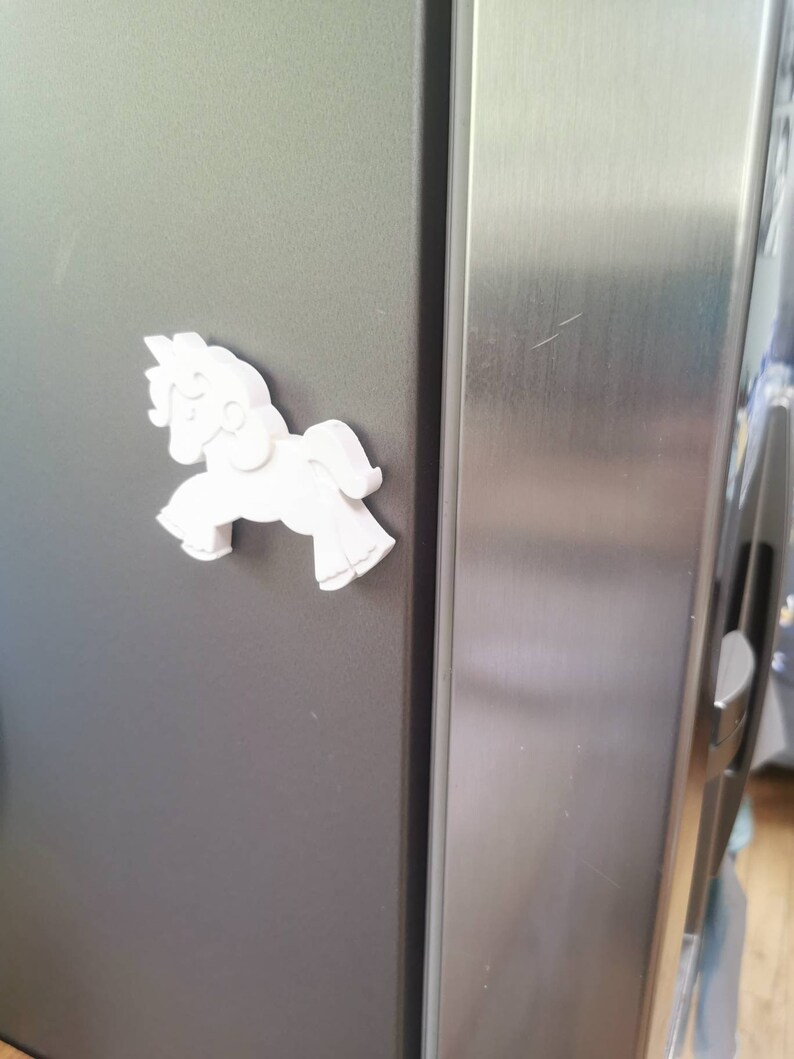 May include: A white, three-dimensional horse-shaped magnet is affixed to a dark gray refrigerator door. The magnet is a stylized depiction of a horse, with a flowing mane and tail. The refrigerator door is next to a stainless steel door.