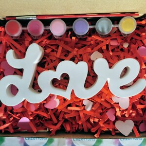 May include: A white wooden "Love" sign with a heart cutout in the "O" sits in a box filled with red shredded paper and pink hearts. The box also contains six small paint pots and a paintbrush.