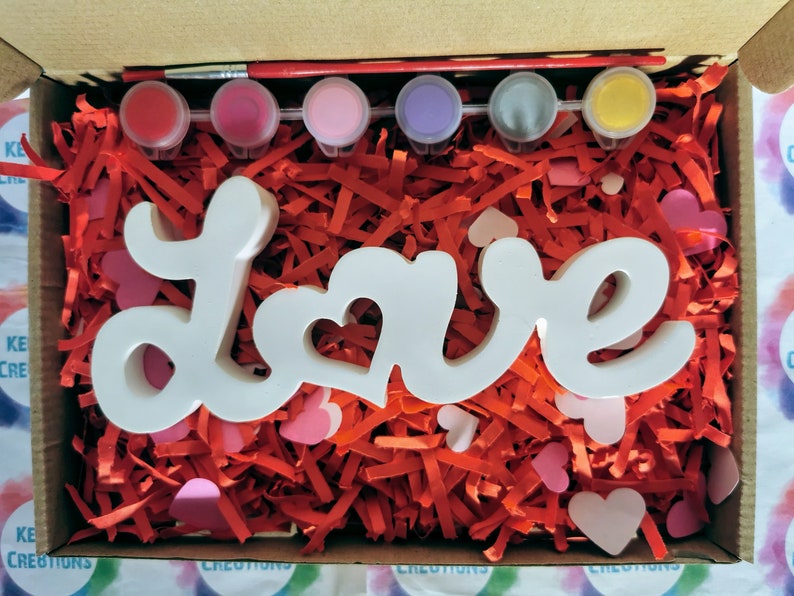 May include: A white wooden "Love" sign with a heart cutout in the "O" sits in a box filled with red shredded paper and pink hearts. The box also contains six small paint pots and a paintbrush.