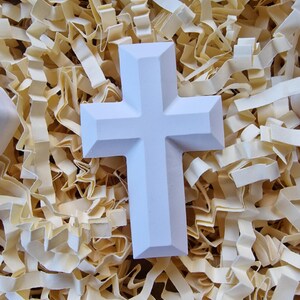 May include: A white ceramic cross with a simple design, resting on a bed of shredded paper.