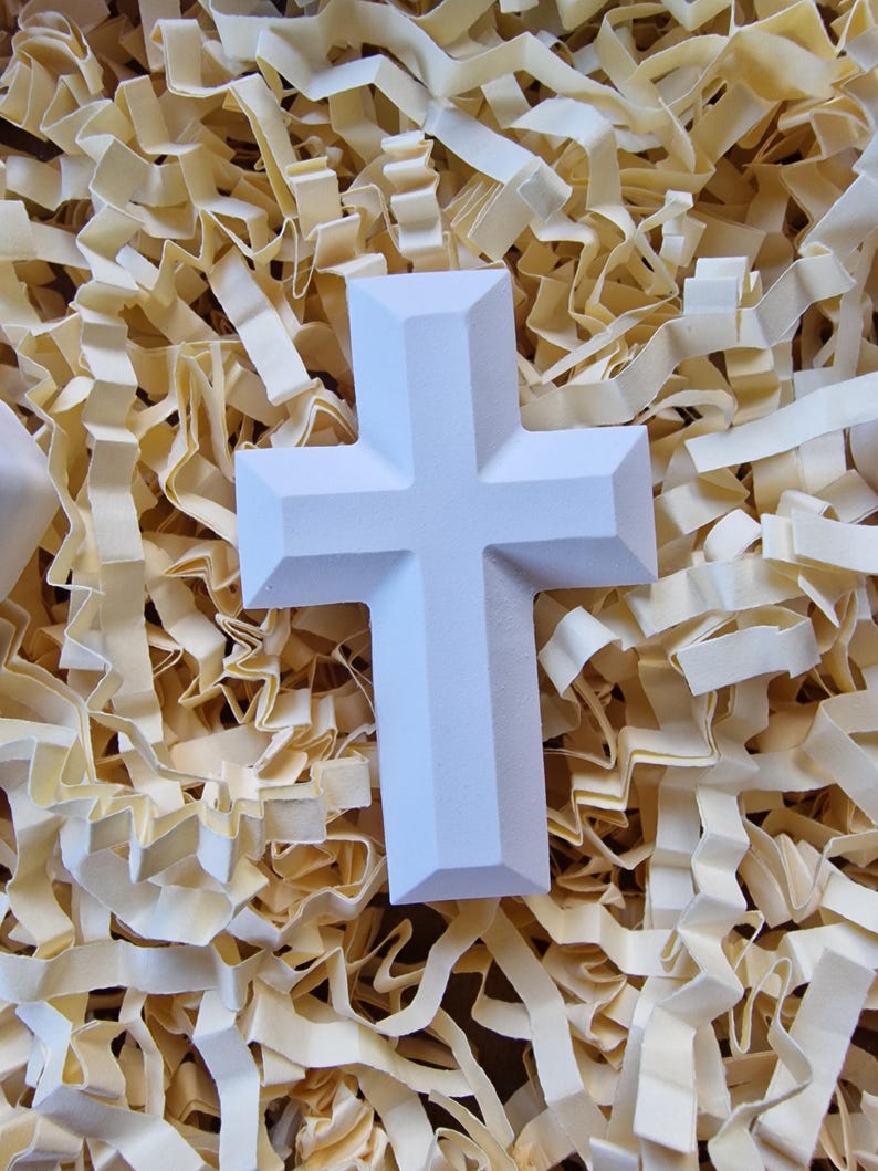 May include: A white ceramic cross with a simple design, resting on a bed of shredded paper.