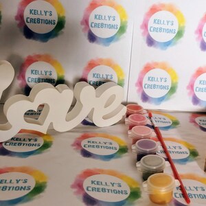 May include: A white wooden "Love" sign with a heart in the "O" is included in a craft kit with paint pots and a brush. The paint pots are pink, red, purple, blue, and gold. The kit is packaged in a brown cardboard box with red shredded paper and pink hearts.