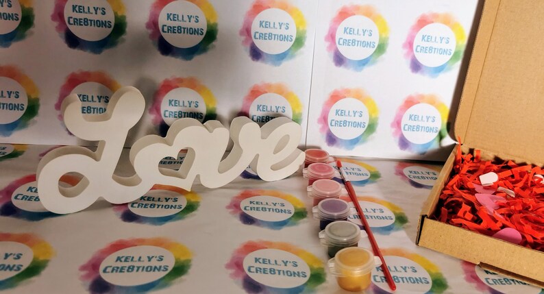 May include: A white wooden "Love" sign with a heart in the "O" is included in a craft kit with paint pots and a brush. The paint pots are pink, red, purple, blue, and gold. The kit is packaged in a brown cardboard box with red shredded paper and pink hearts.