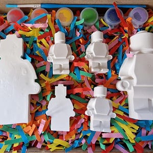 May include: A set of five white ceramic figurines shaped like Lego people, with paint pots and a paintbrush. The figurines are sitting on a bed of colorful shredded paper.