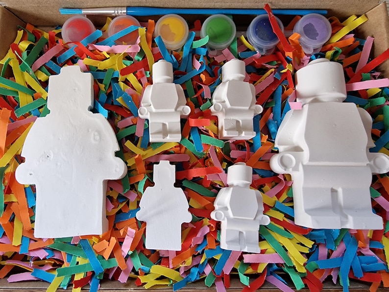 May include: A set of five white ceramic figurines shaped like Lego people, with paint pots and a paintbrush. The figurines are sitting on a bed of colorful shredded paper.