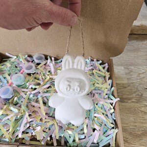 Peut inclure: Ornement blanc en forme de lapin, avec une ficelle pour suspendre, tenu par une main. L'ornement est &agrave; l'int&eacute;rieur d'une bo&icirc;te en carton remplie de papier d&eacute;chiquet&eacute; color&eacute;. Petits pots de peinture visibles.