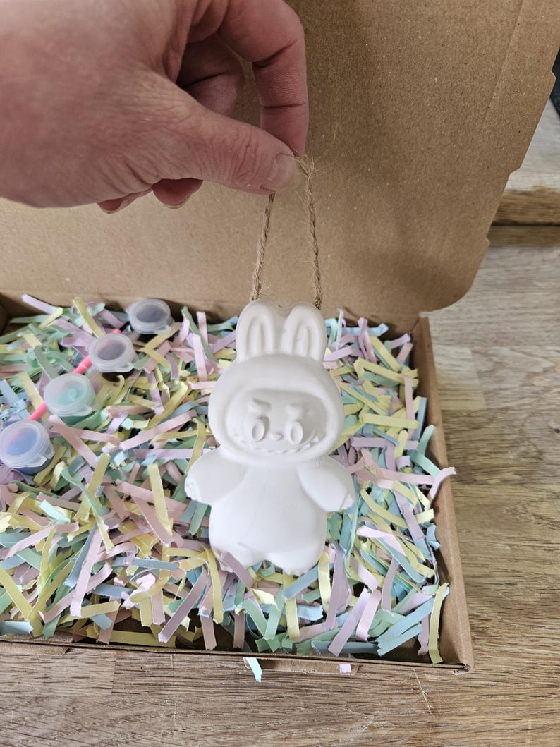 Peut inclure: Ornement blanc en forme de lapin, avec une ficelle pour suspendre, tenu par une main. L'ornement est &agrave; l'int&eacute;rieur d'une bo&icirc;te en carton remplie de papier d&eacute;chiquet&eacute; color&eacute;. Petits pots de peinture visibles.