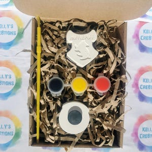 May include: A craft kit in a cardboard box, featuring a Hufflepuff crest, paint pots in black, yellow, and red, a paint brush, and a circular object. The box is filled with brown paper shreds. The text "KELLY'S CREATIONS" is visible on the surrounding stickers.
