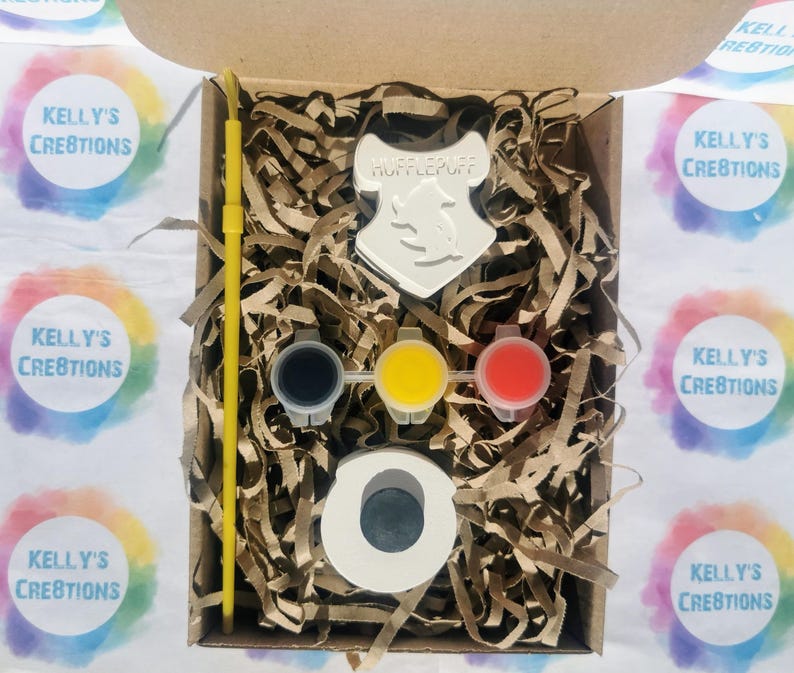 May include: A craft kit in a cardboard box, featuring a Hufflepuff crest, paint pots in black, yellow, and red, a paint brush, and a circular object. The box is filled with brown paper shreds. The text "KELLY'S CREATIONS" is visible on the surrounding stickers.