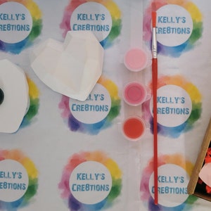 May include: A craft kit for painting a heart-shaped ceramic ornament. The kit includes three small pots of paint in shades of pink and red, a paintbrush, and two heart-shaped ceramic ornaments. The ornaments are white and have a textured surface. The kit is packaged in a box with a colorful background and the text "Kelly's Creations" printed on it.