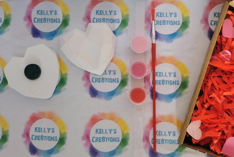 May include: A craft kit for painting a heart-shaped ceramic ornament. The kit includes three small pots of paint in shades of pink and red, a paintbrush, and two heart-shaped ceramic ornaments. The ornaments are white and have a textured surface. The kit is packaged in a box with a colorful background and the text "Kelly's Creations" printed on it.