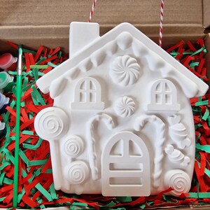 May include: A white ceramic gingerbread house ornament with candy cane, lollipop, and swirl designs. The ornament is in a box with red and green shredded paper and a paintbrush and paint pots.