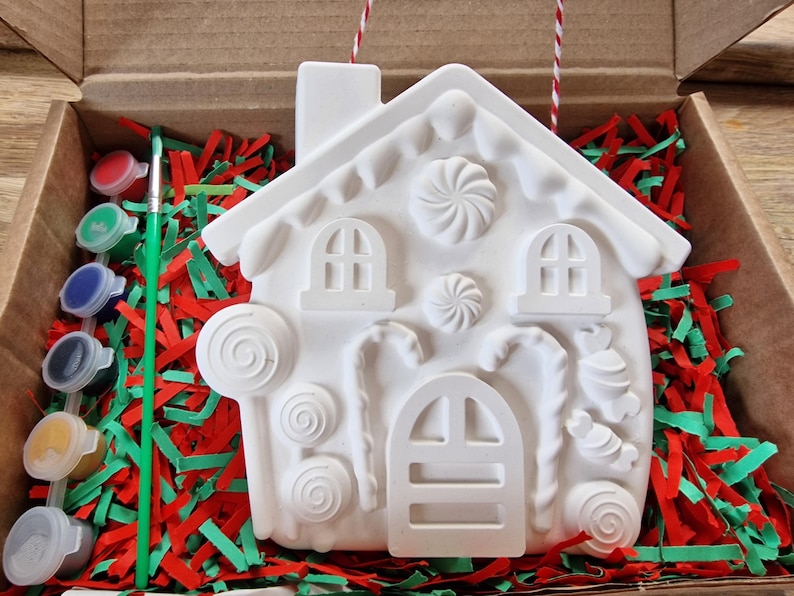 May include: A white ceramic gingerbread house ornament with candy cane, lollipop, and swirl designs. The ornament is in a box with red and green shredded paper and a paintbrush and paint pots.
