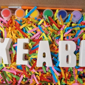 Op de afbeelding: Een set witte keramische letters die de naam "KEABA" spellen in een doos gevuld met kleurrijk versnipperd papier. De letters zijn klaar om te worden geschilderd met de meegeleverde set van zes kleine verfpotjes in rood, oranje, geel, groen, blauw en paars. Een kwast is ook inbegrepen.