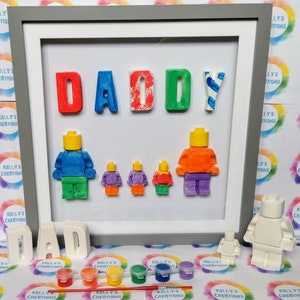 May include: A framed craft project with the word "DADDY" spelled out in colorful letters. There are five small, unpainted figurines that look like Lego people, and one larger unpainted figurine that looks like a Lego person. There are also small paint pots and a paintbrush.