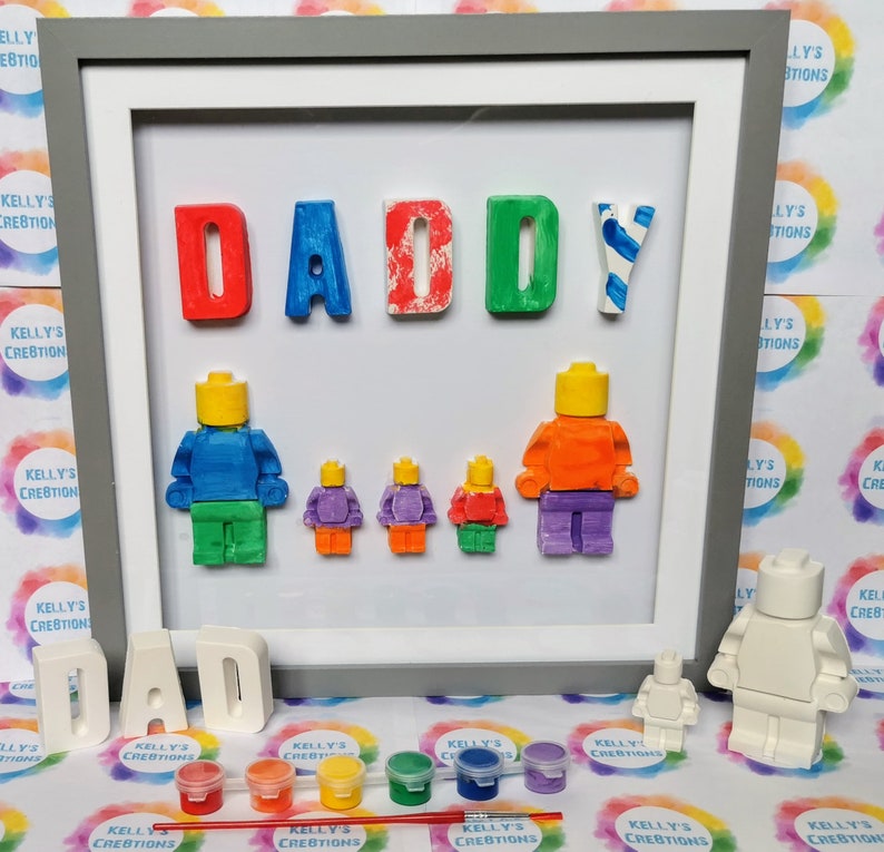 May include: A framed craft project with the word "DADDY" spelled out in colorful letters. There are five small, unpainted figurines that look like Lego people, and one larger unpainted figurine that looks like a Lego person. There are also small paint pots and a paintbrush.