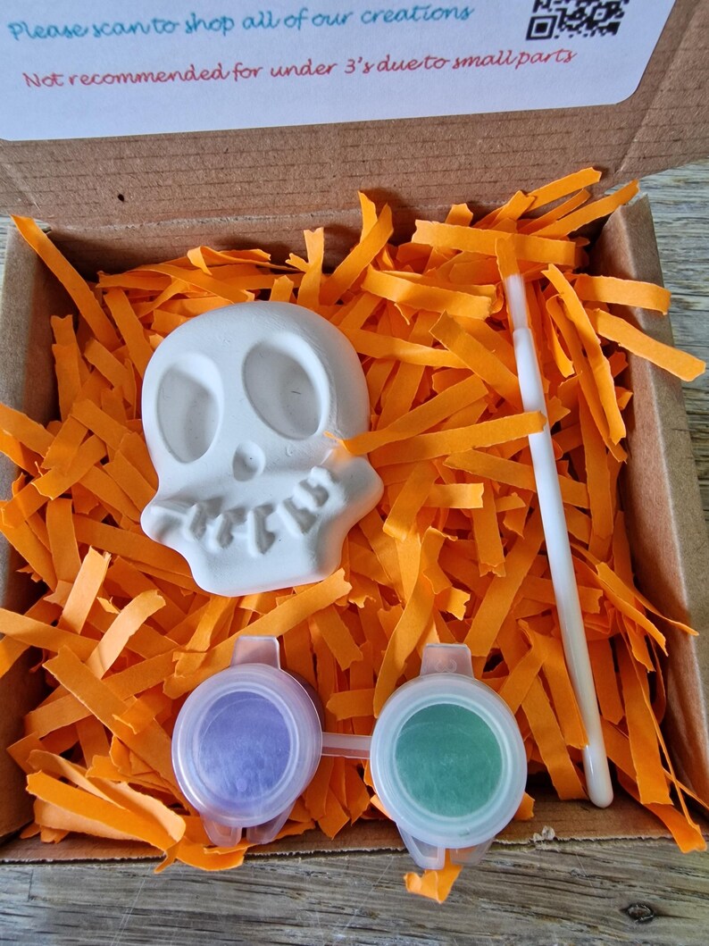 May include: A craft kit in a cardboard box, featuring a white skull, two paint pots with purple and green paint, and a paintbrush. The box is filled with orange paper strips. Text on the box reads: "Please scan to shop all of our creations" and "Not recommended for under 3's due to small parts."