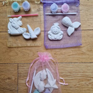 Puede incluir: Tres bolsas de organza llenas de pintura, pinceles y figuritas de cer&aacute;mica para pintar. Las bolsas son amarillas, moradas y rosas. Las figuritas incluyen un unicornio, una sirena, una princesa, un arco&iacute;ris y una estrella.
